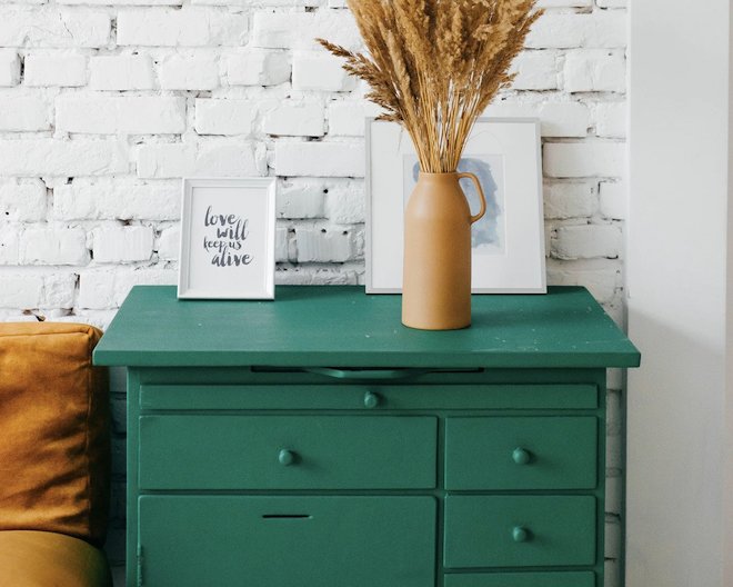 image of a teal chest of drawers with a white brick background, a framed note that says 'love will keep us alive', and a terracotta vase with wheat stalks in it